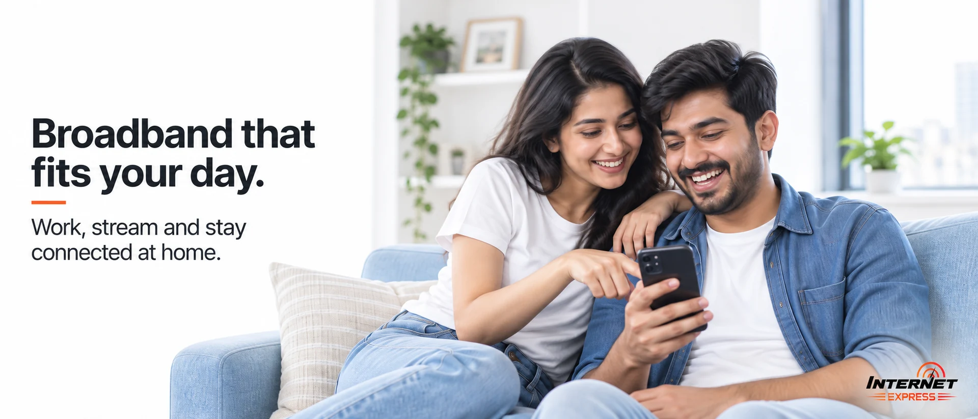 Young couple sitting together on a sofa at home, looking at a phone. Caption: broadband that fits your day — work, stream and stay connected at home.