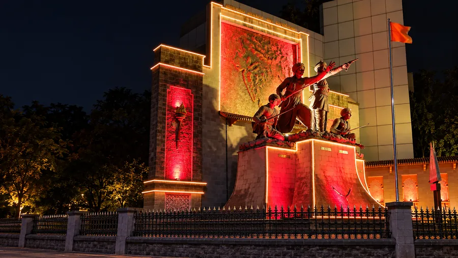 Floodlit stone memorial at night showing a sculpted group of figures on a plinth in front of a carved relief panel, edged with warm light strips, with a saffron flag on a pole to one side.