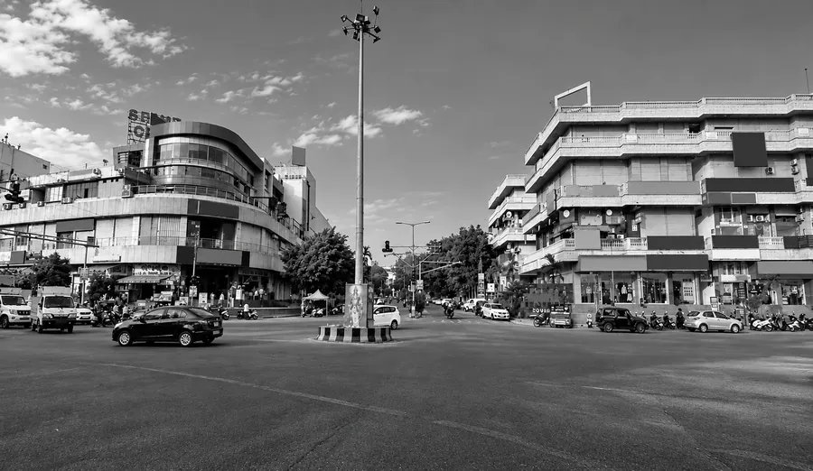 Wide road junction with a curved multi-storey commercial building on one side, a taller residential and shop block opposite, and a tall lighting mast on the island between them.