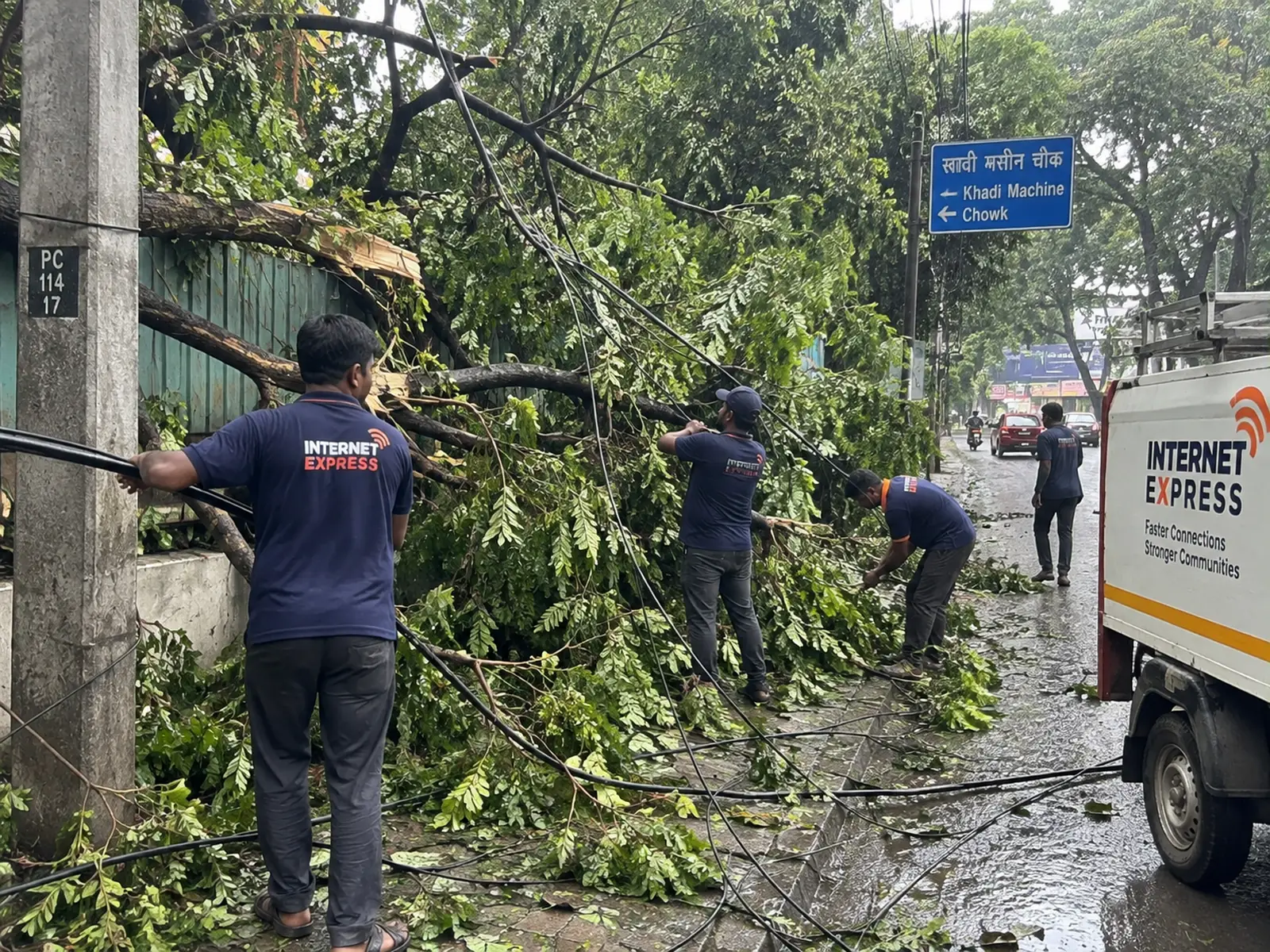 An Internet Express crew clearing a fallen tree from telecom cables at Khadi Machine Chowk, Pune, with the branch resting across the aerial span and the team van alongside.
