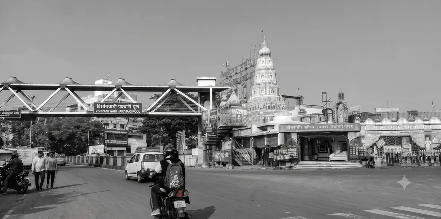 Street view at Vishrantwadi with a steel pedestrian footbridge carrying a Vishrantwadi Padchari Pool nameplate, a tall ornate temple spire behind it and traffic crossing the junction.